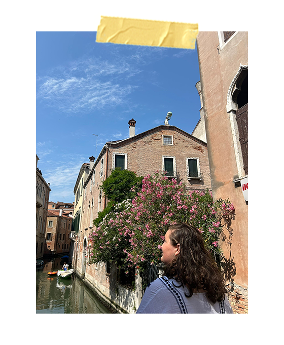 A photo of me looking off of a smaller bridge in Venice with the river and pretty pink flowers in the background.